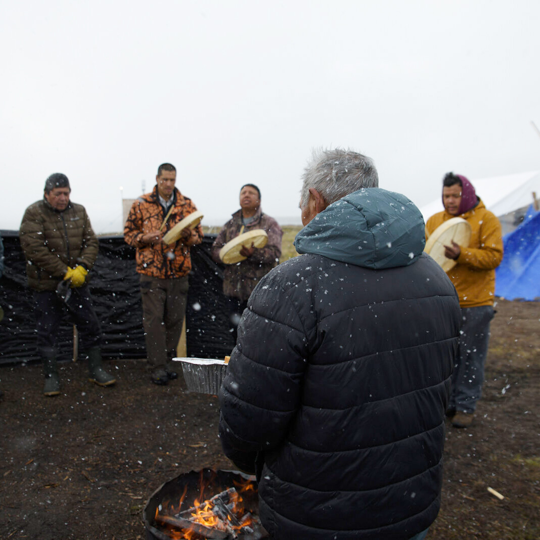 Drummers sing while Louis Zoe feeds the fire with an offering of bread and tobacco, a way to ask The Creator for safe travel and offer thanks before the day begins.